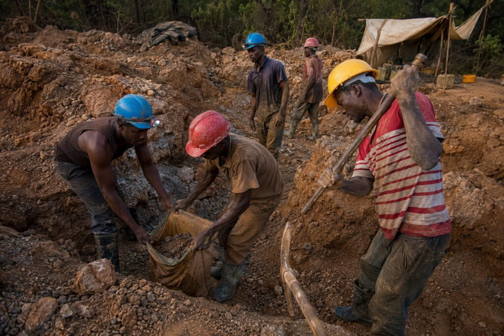 Artisanal miners digging and shovelling ore by hand at a dusty open pit mining site in Zambia, wearing mixed safety gear including hard hats