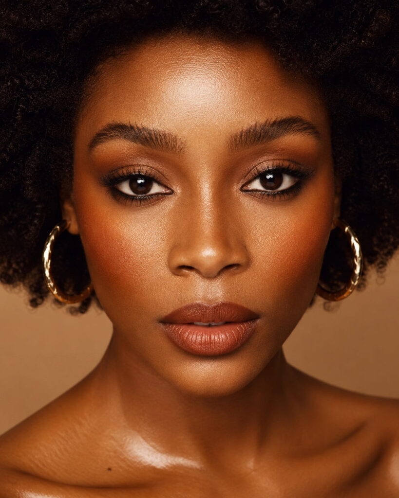 Close-up beauty portrait of a Black woman with warm amber eyeshadow, smudged brown liner, terracotta lip and peachy bronze blush — 1970s makeup and beauty trends for women