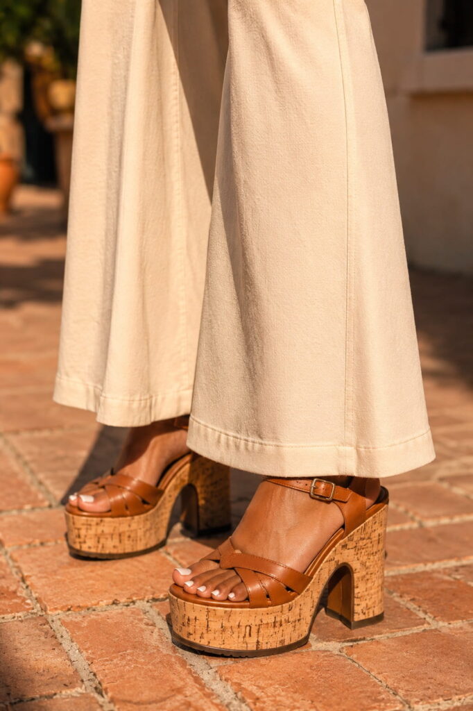 Close-up of cream wide-leg flared trousers grazing tan cork platform sandals with ankle strap on a warm tiled floor — 1970s platform shoes women 70s outfit styling detail